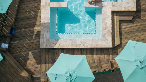Een rechthoekig zwembad met een houten terras eromheen, inclusief een kleine waterval en een zitgedeelte met parasols.