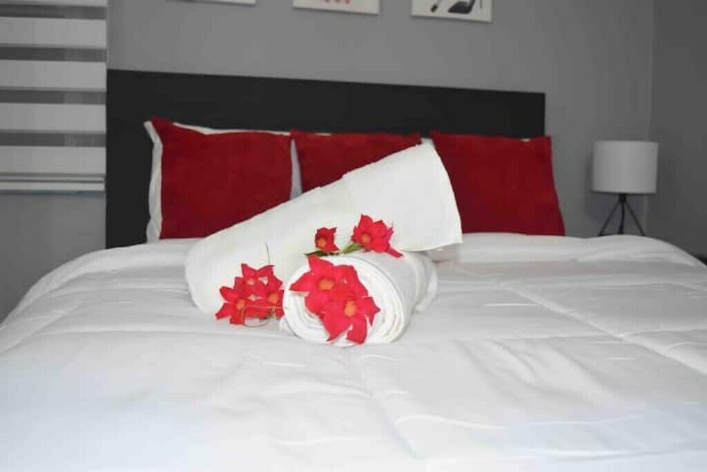 A neatly made bed with white bedding and red pillows, a rolled white towel with red flowers, and a lamp on a side table.