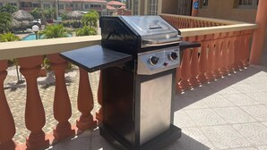A barbecue grill on an outdoor balcony with a pool and buildings in the background.