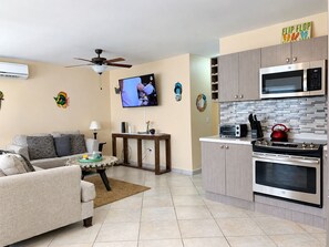 A modern living room with a sofa, a coffee table, and a TV. There's a kitchen area with a microwave, oven, and a kettle.