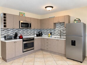 A modern kitchen with light wood cabinets, a stainless steel refrigerator, a microwave, a toaster, and a kettle.