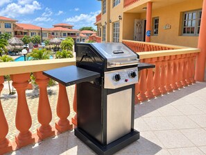A barbecue grill on an outdoor balcony with a pool and hotel buildings in the background.
