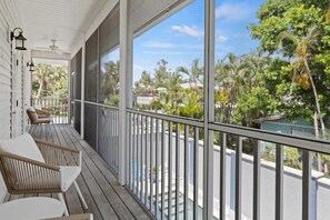 A covered porch with wicker chairs, a ceiling fan, and large windows overlooking a street with palm trees. - Stunning Coastal Home with Pool, Steps to Beach (Fort Myers Beach)