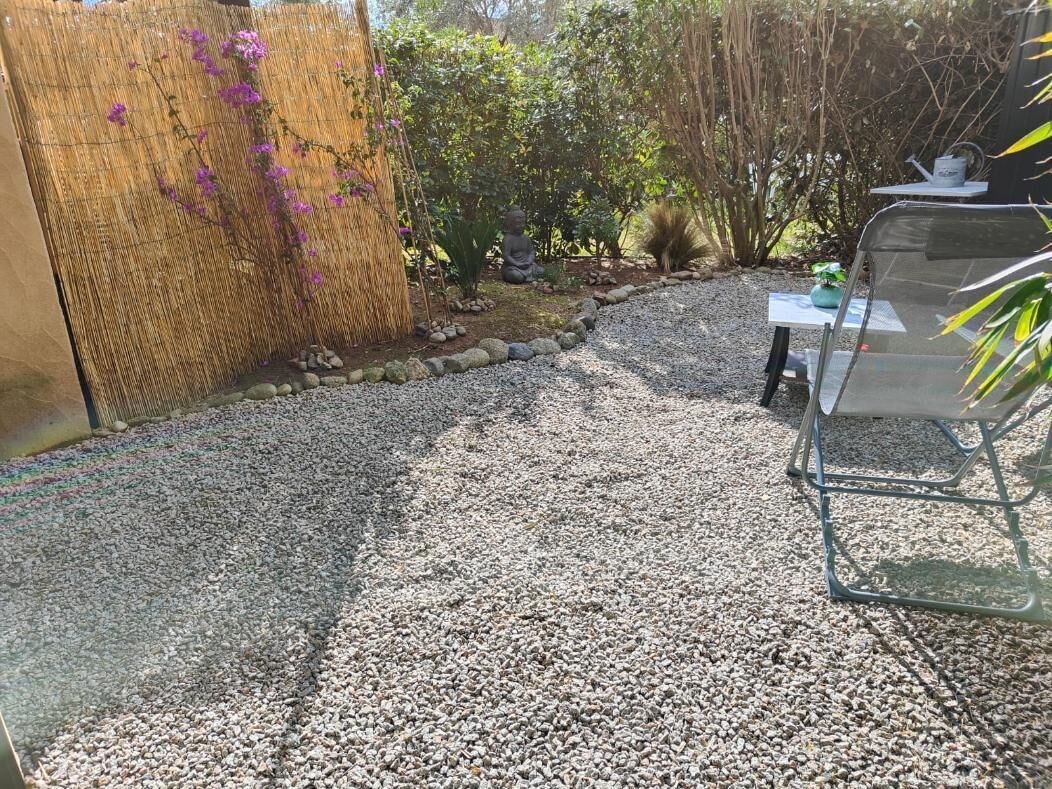 A gravel driveway leading to a covered patio area with a seating arrangement and a small garden with a Buddha statue.