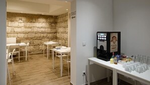 Una habitación con pared de piedra, una mesa blanca con sillas, una cafetera y un letrero indicando el baño.