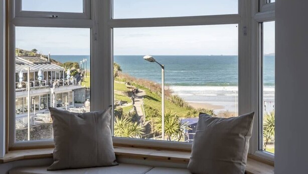 Vista desde una ventana con dos almohadas, con vista a una playa y una carretera costera con edificios y vegetación.