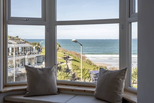 Une vue depuis une fenêtre, avec deux coussins, donnant sur une plage et une route côtière bordée de bâtiments et de verdure.
