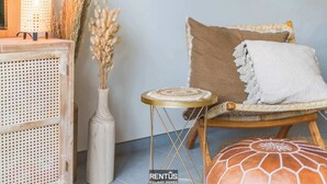 Un rincón acogedor con una silla de mimbre, una mesita redonda pequeña, un jarrón blanco con plantas secas y un aparador de madera rústica.