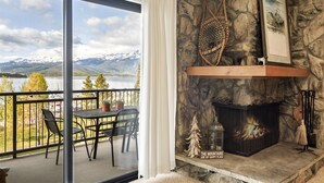 A balcony with a table and chairs, a fireplace with a stone wall backdrop, and a scenic view of mountains and a lake.