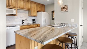 A modern kitchen with wooden cabinets, a marble countertop, a built-in oven, and a refrigerator. There are two bar stools and pendant lights above the island.