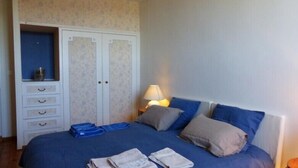 Ein Schlafzimmer mit Bett, Kissen, einer Lampe, einem Kleiderschrank und gefalteter Kleidung auf dem Bett.