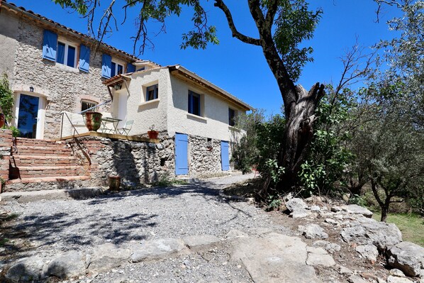 A stone building with blue shutters, a gravel path, and a large tree.