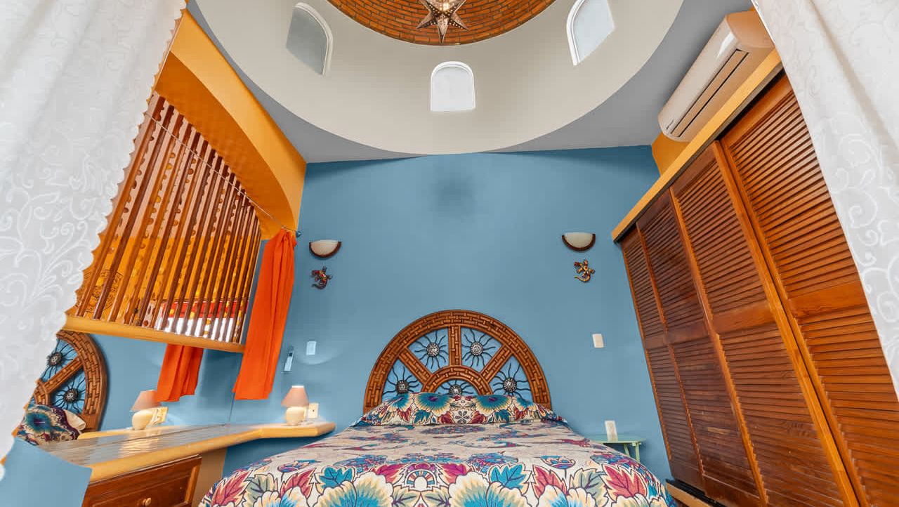 Une chambre à coucher avec un plafond incurvé, un lit doté d’une tête de lit à motifs, une table de chevet, une commode et une fenêtre avec des stores.