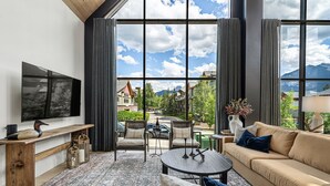 A modern living room with a large TV, comfortable seating, and a view of a mountain landscape. - Spectacular 8-BDRM, 8-BATH Mountain Retreat! (Canmore)