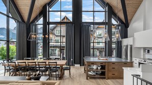 A spacious living area with a wooden ceiling, large windows, and a kitchen with modern appliances. - Spectacular 8-BDRM, 8-BATH Mountain Retreat! (Canmore)