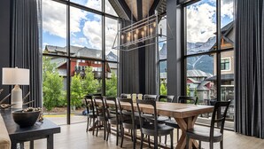 A dining area with a large table and chairs, a hanging light fixture, and a view of mountains through large windows. - Spectacular 8-BDRM, 8-BATH Mountain Retreat! (Canmore)