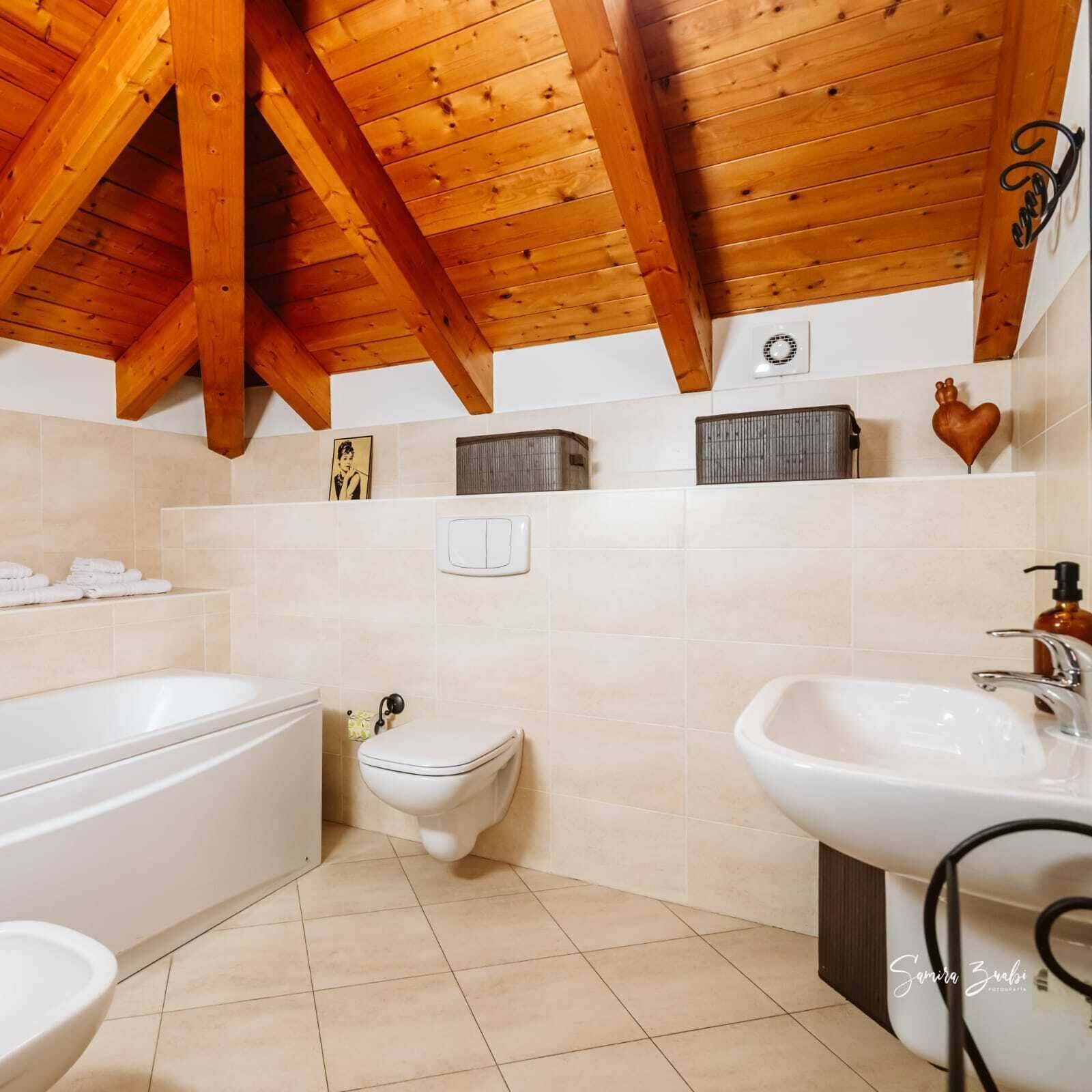 A bathroom with a bathtub, toilet, and sink, wooden ceiling beams, and a tiled floor.