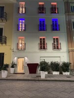 Un edificio de varios pisos con balcones, un toldo rojo y plantas en macetas en la entrada.