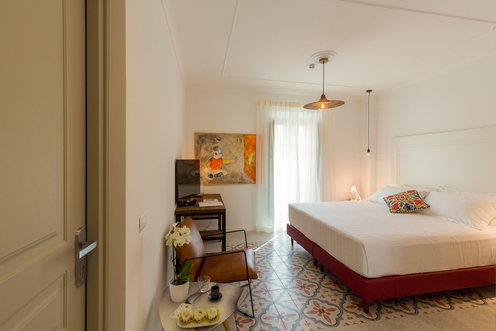 A hotel room with a bed, a desk, a chair, a small table with a plant and a tray of food, and a painting on the wall.