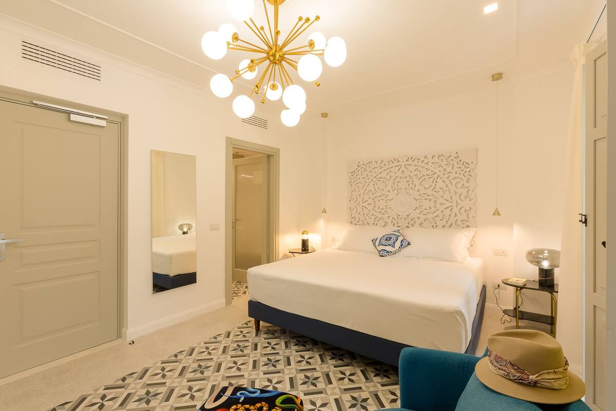 A modern bedroom with a large bed, a decorative wall panel, a unique chandelier, and a patterned rug.