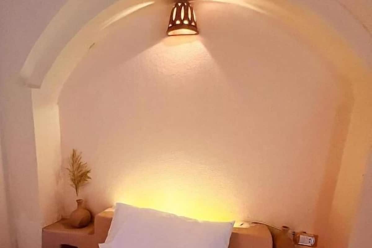 Une chambre avec un lit simple, une table de chevet, un luminaire fixé au mur et un petit vase contenant des plantes séchées.