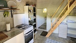Eine kleine Küche mit Gasherd, Backofen und weißen Schränken. Eine Holztreppe führt zu einem Schlafzimmer mit Bett und Kommode.