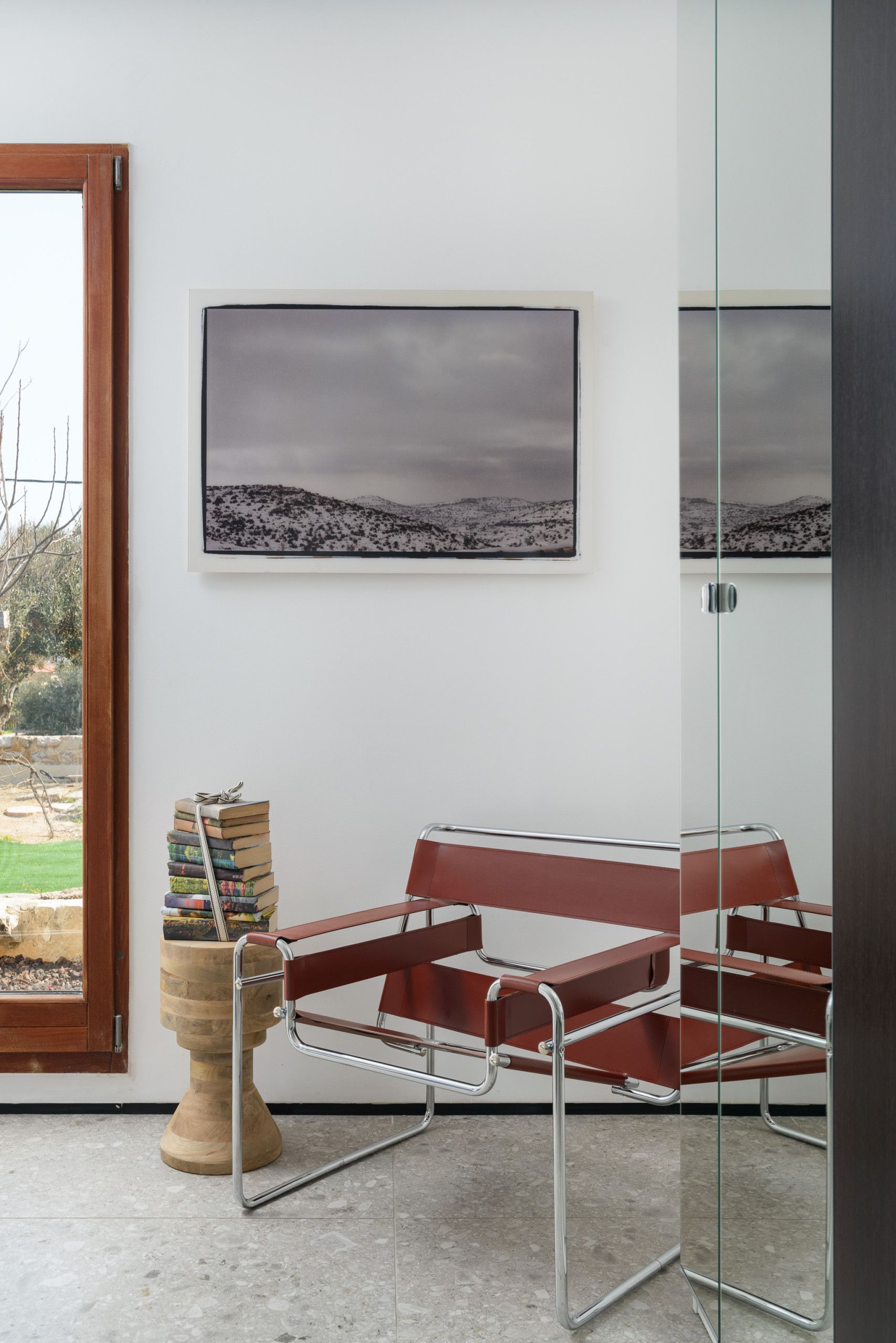 Una habitación moderna con puerta de vidrio, una silla roja, un taburete de madera con libros y un cuadro con paisaje en la pared.