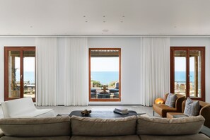 A modern living room with a sofa, armchair, and a coffee table. There are large windows with curtains, offering a view of the sea. - Luxury Villa over the Cliffs by FeelHome (Hefer Valley)