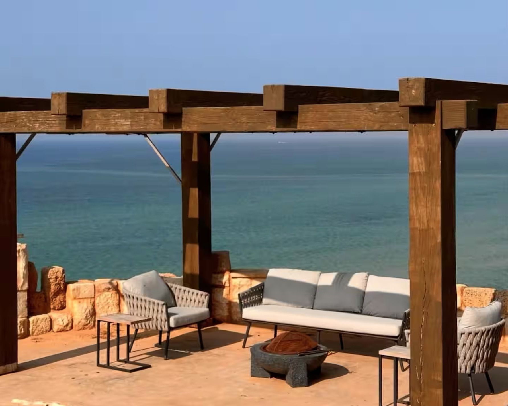 A wooden pergola with a white sofa and two armchairs, overlooking a calm sea.