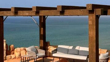 A wooden pergola with a white sofa and two armchairs, overlooking a calm sea.