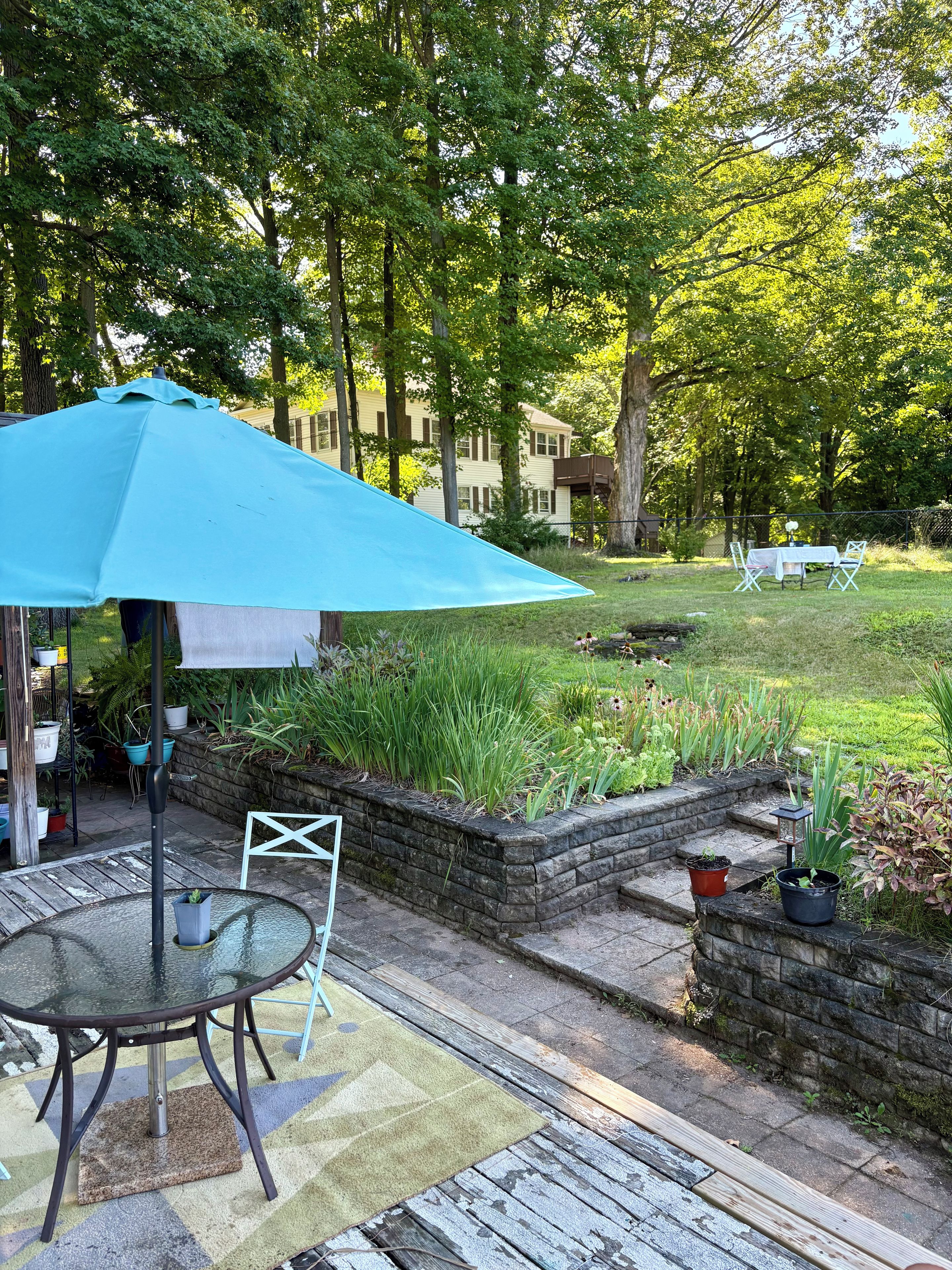 A patio with a table and chairs, a blue umbrella, and a garden with plants and a brick wall.
