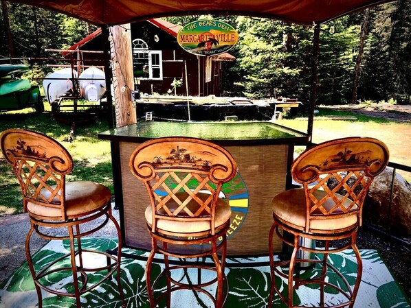Una barra de madera con tres sillas, un letrero que dice 'The Bears Den Margaritaville' y una casa al fondo.