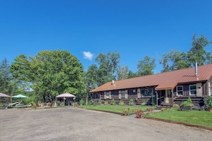 A rustic log cabin with a metal roof, surrounded by a gravel driveway and lush greenery. - Unique and charming, exquisitely themed Log Cabin (Hamilton)