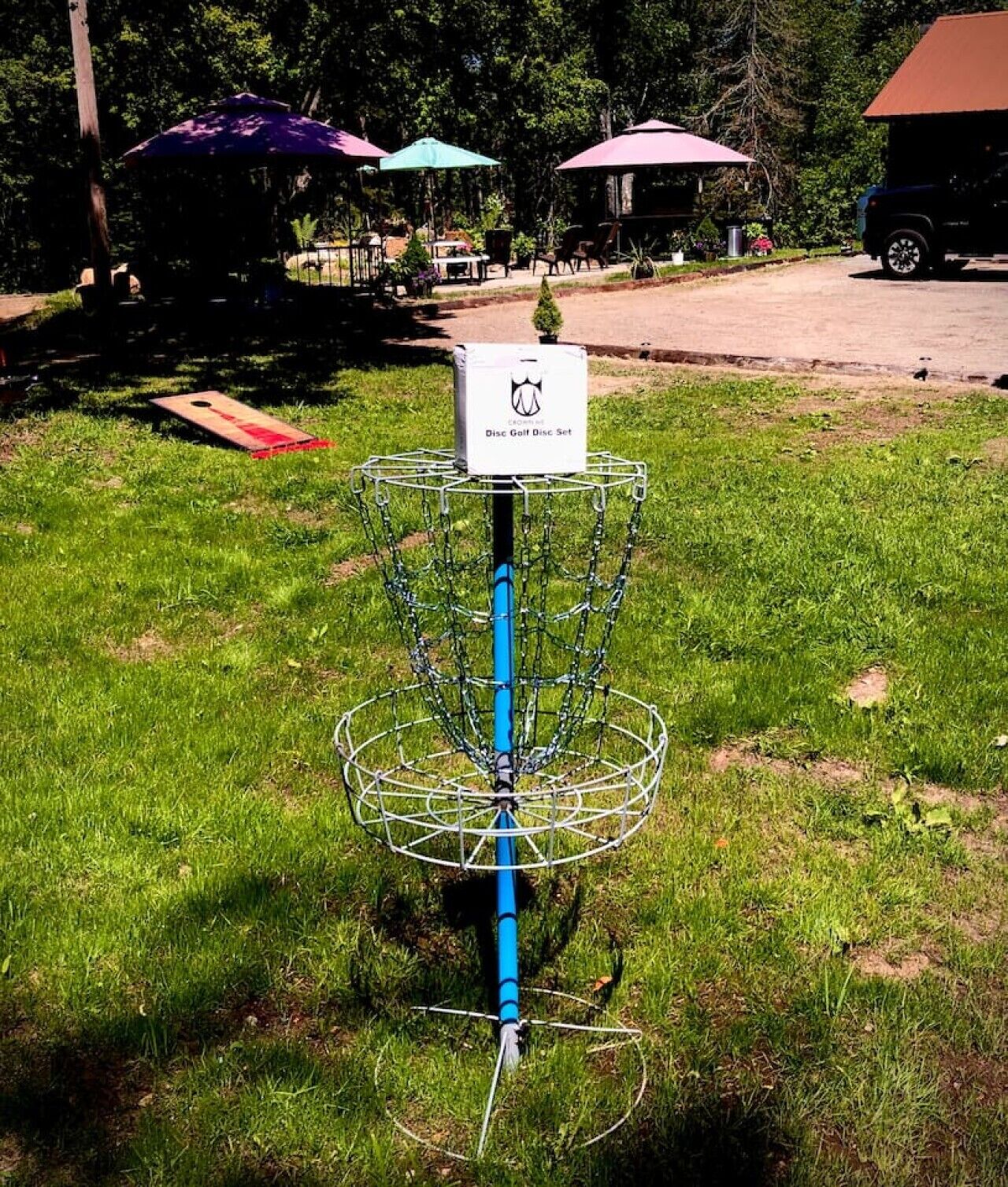 A disc golf basket with a chain cage, a disc golf disc set box, and a parking area with umbrellas in the background.