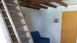 Een houten trap die leidt naar een hoger gelegen niveau, een blauwe bank en een houten deur in een kamer met witte muren.