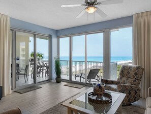 Relax in this inviting living area with a balcony overlooking the beach, featuring a ceiling fan, coffee table, and comfortable armchair.