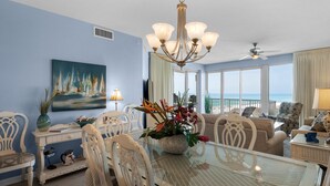 A spacious living room with a glass-top dining table, white chairs, a chandelier, and a painting of sailboats. - Beautiful Gulf Front 2 Bedroom Condo in Destin (Miramar Beach)