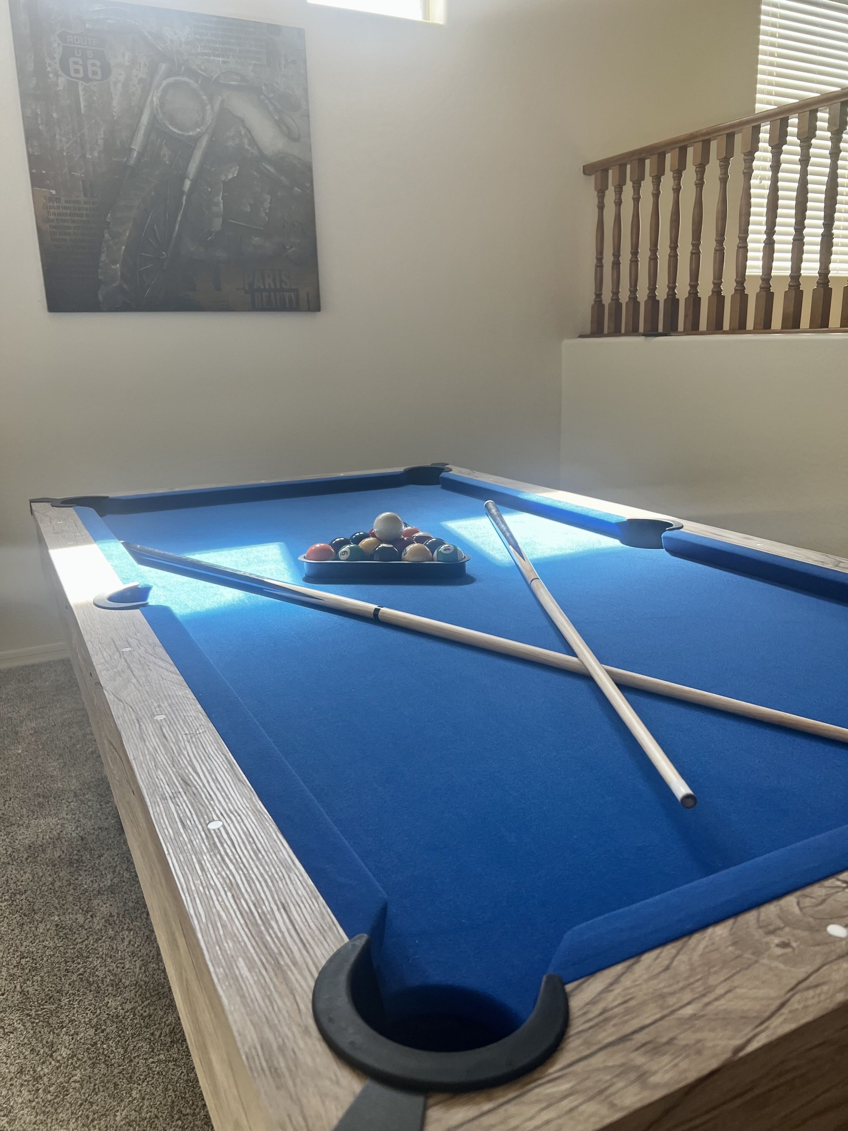 Una mesa de billar con superficie de fieltro azul, tacos y un cesto de bolas.