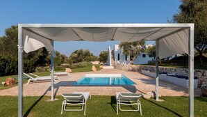 A covered outdoor swimming pool with two lounge chairs, a stone wall, and a white building in the background. - Trullo Delle Stelle by Wonderful Italy (Ostuni)