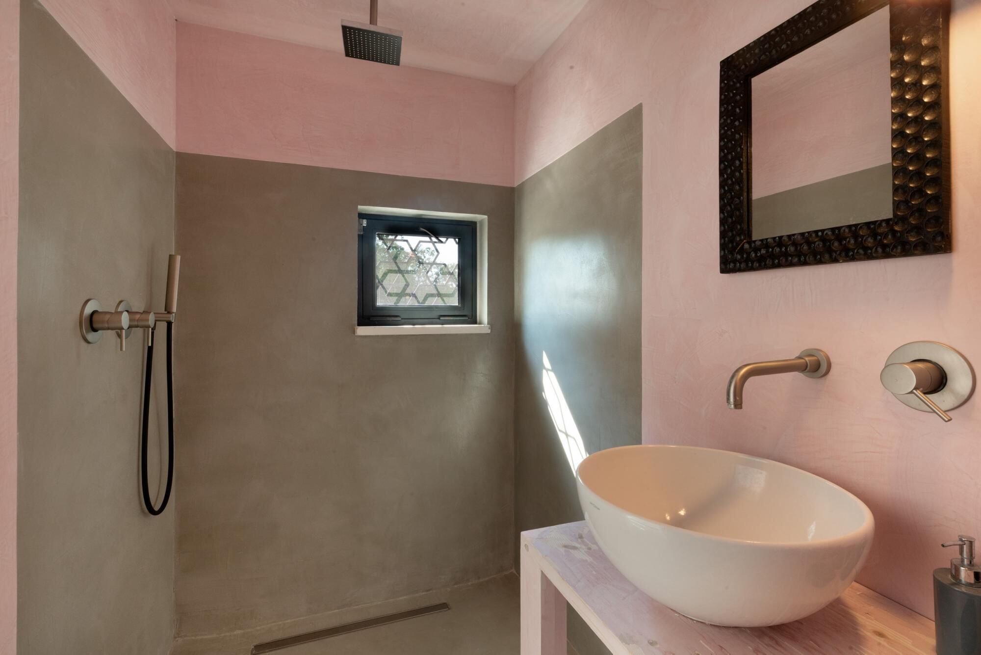 A bathroom with a large white sink, a shower with a handheld showerhead, a small window, and a mirror with decorative lights.