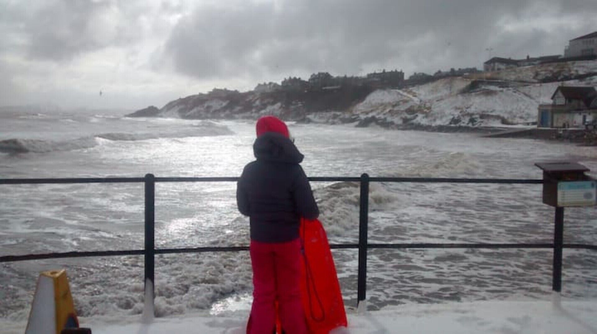 Una persona in piedi accanto a una ringhiera che si affaccia su un mare agitato, con una cittadina costiera sullo sfondo.