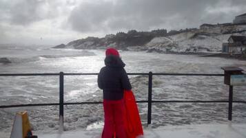 Una persona in piedi accanto a una ringhiera che si affaccia su un mare agitato, con una cittadina costiera sullo sfondo.
