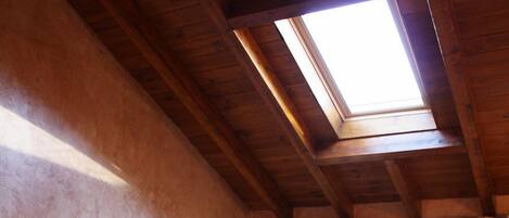 A bedroom with a skylight, wooden ceiling beams, a bed with pillows, a nightstand, and curtains.