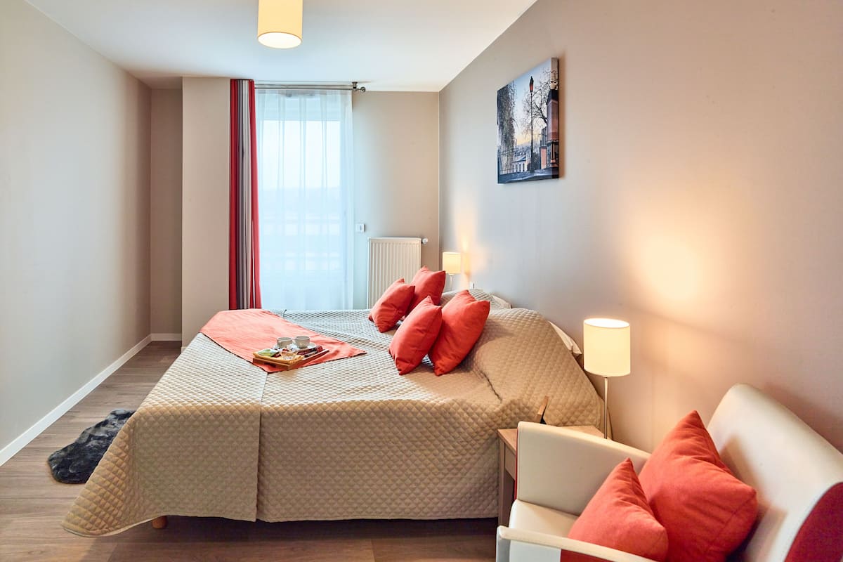 A bedroom with a bed, a chair, a lamp, a painting, and a window with curtains.