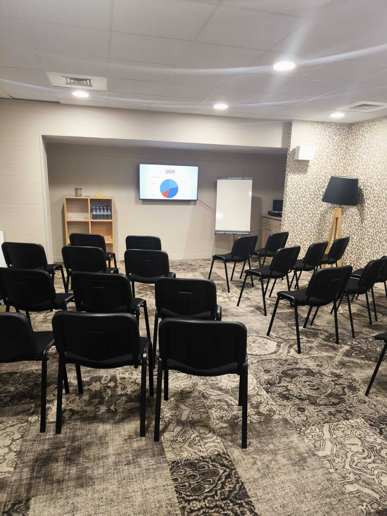 A conference room with rows of black chairs, a carpeted floor, a whiteboard, a screen displaying a pie chart, and a shelf with bottled water.