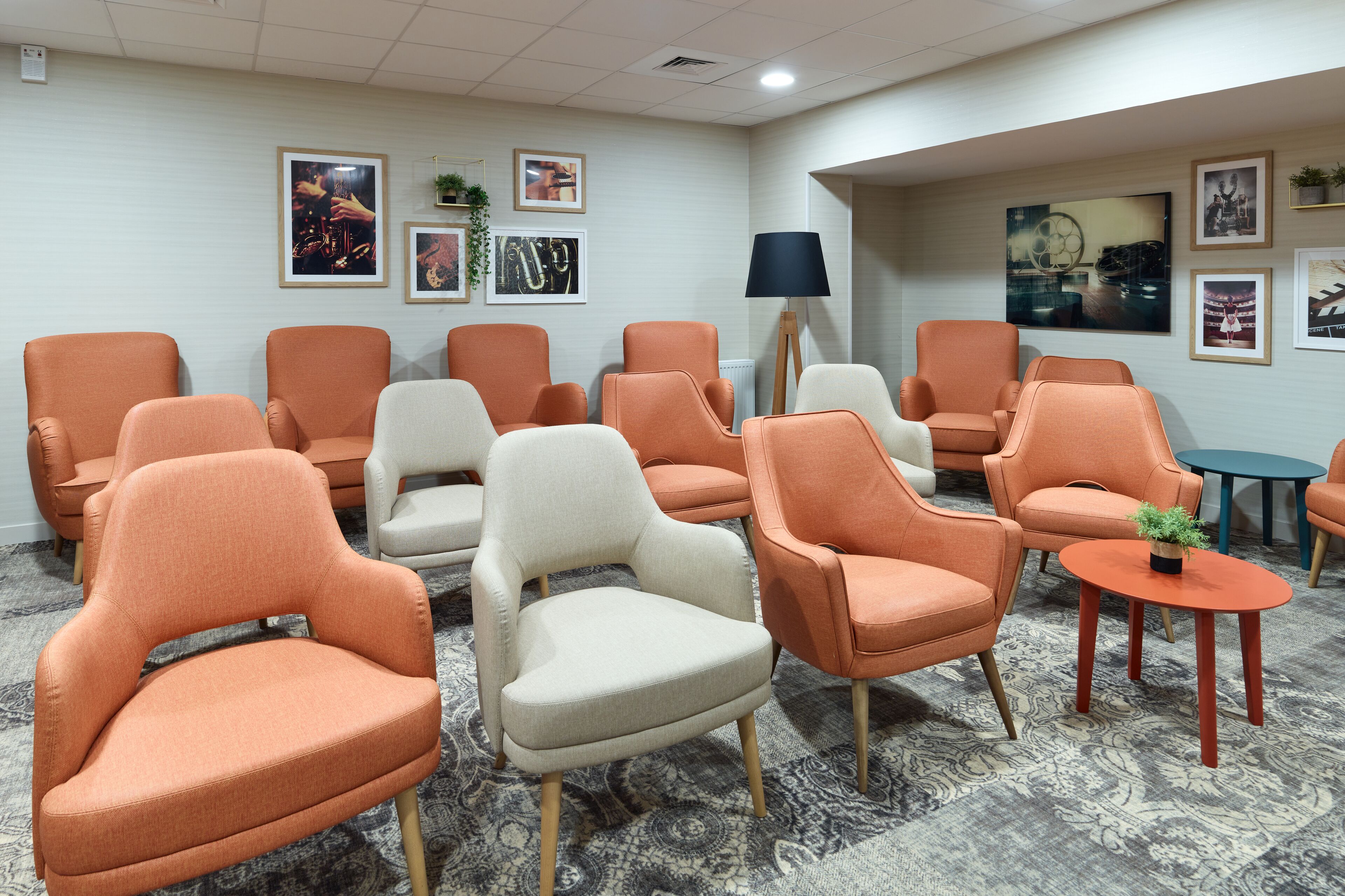 A waiting area with multiple orange and white chairs arranged around a central table, a floor lamp, and framed artwork on the walls.
