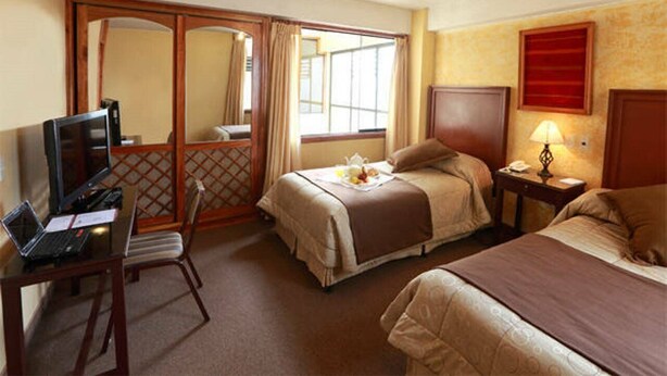 A hotel room with two beds, a desk with a laptop, a TV, and a window with curtains. - El Puma (Cusco)