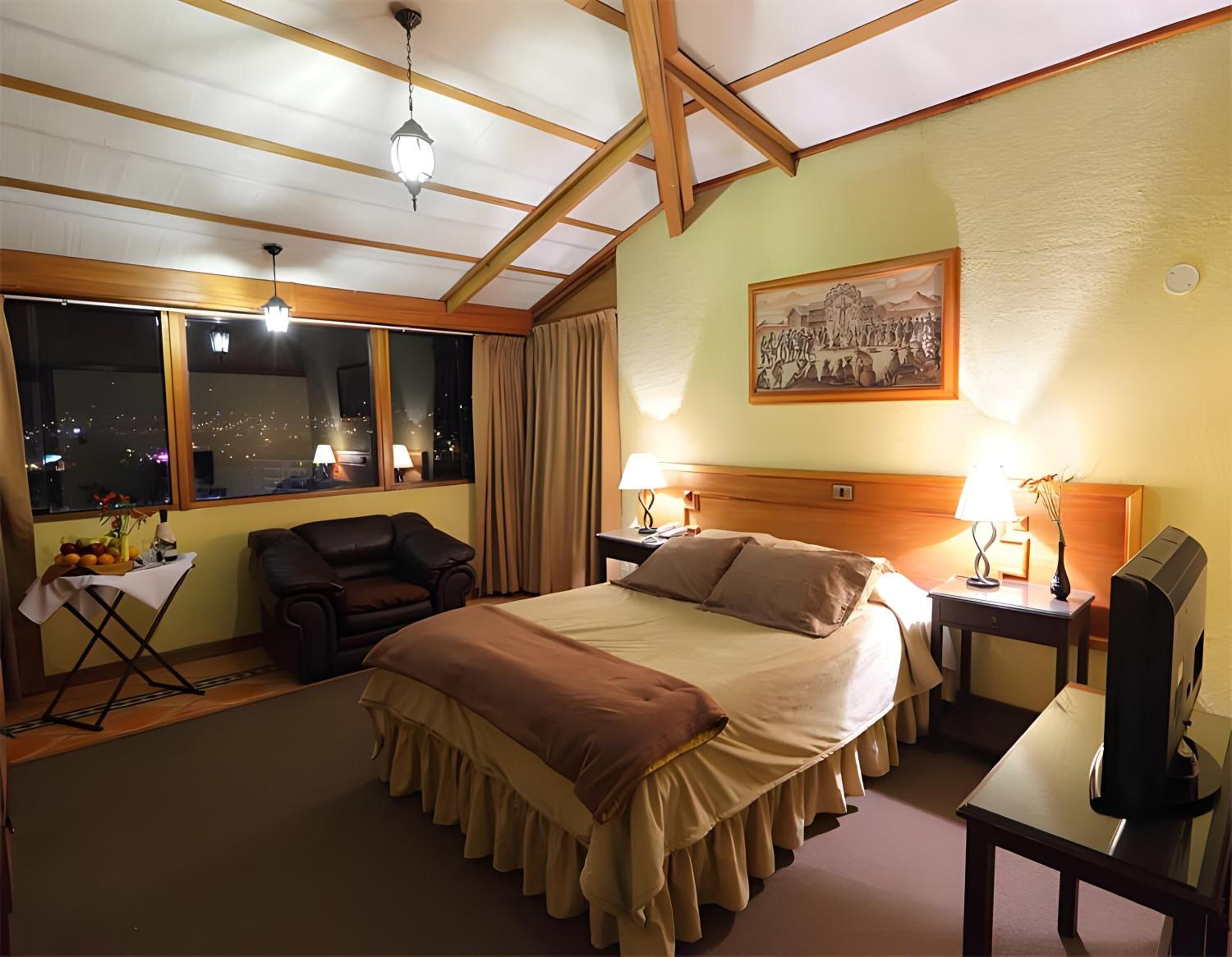 Quarto de hotel com uma cama grande, uma escrivaninha, uma cadeira, uma janela com vista para a cidade e um quadro na parede.