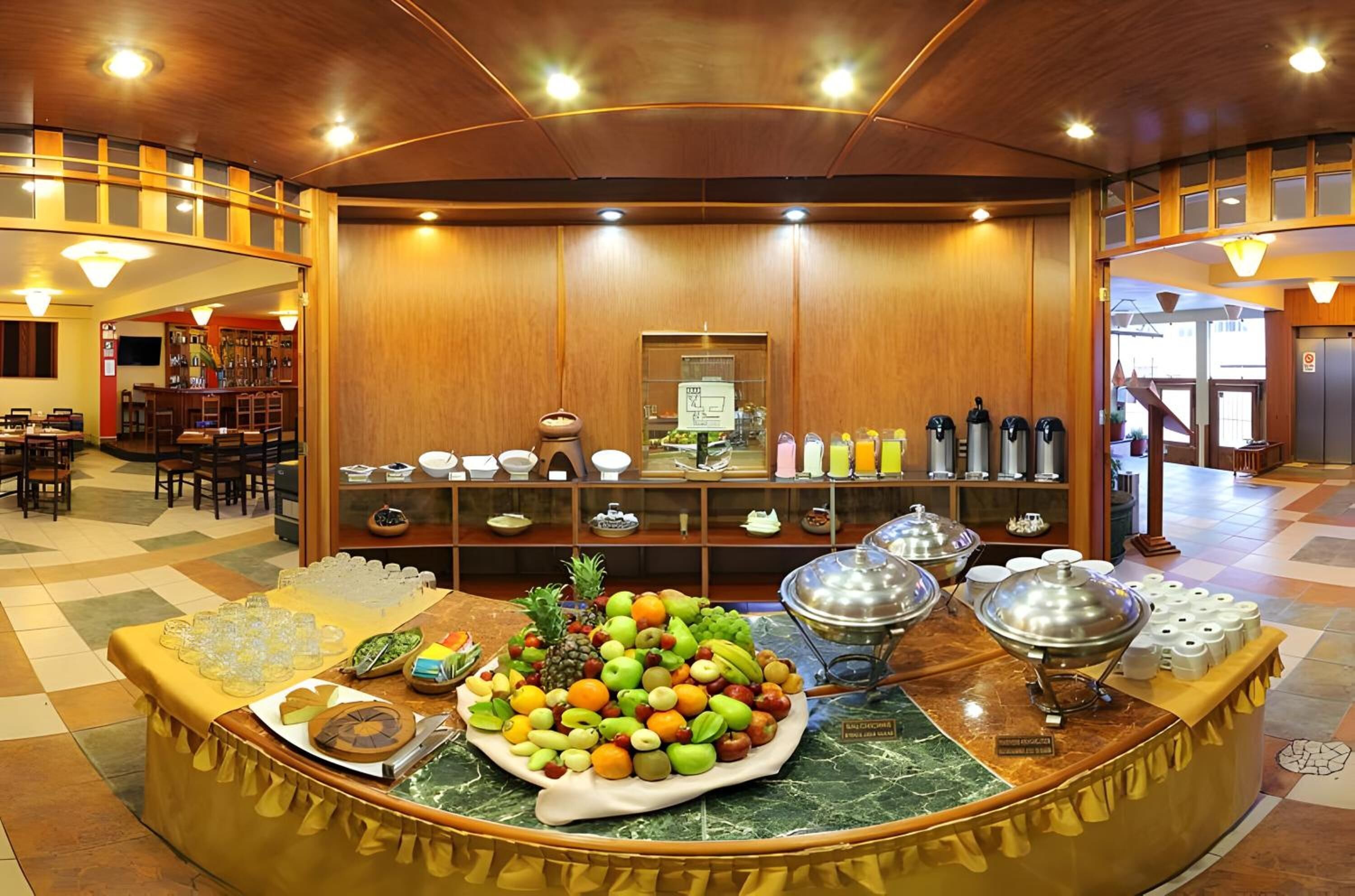 Uma área de buffet com diversas frutas e bebidas, painéis de madeira e um balcão de recepção.