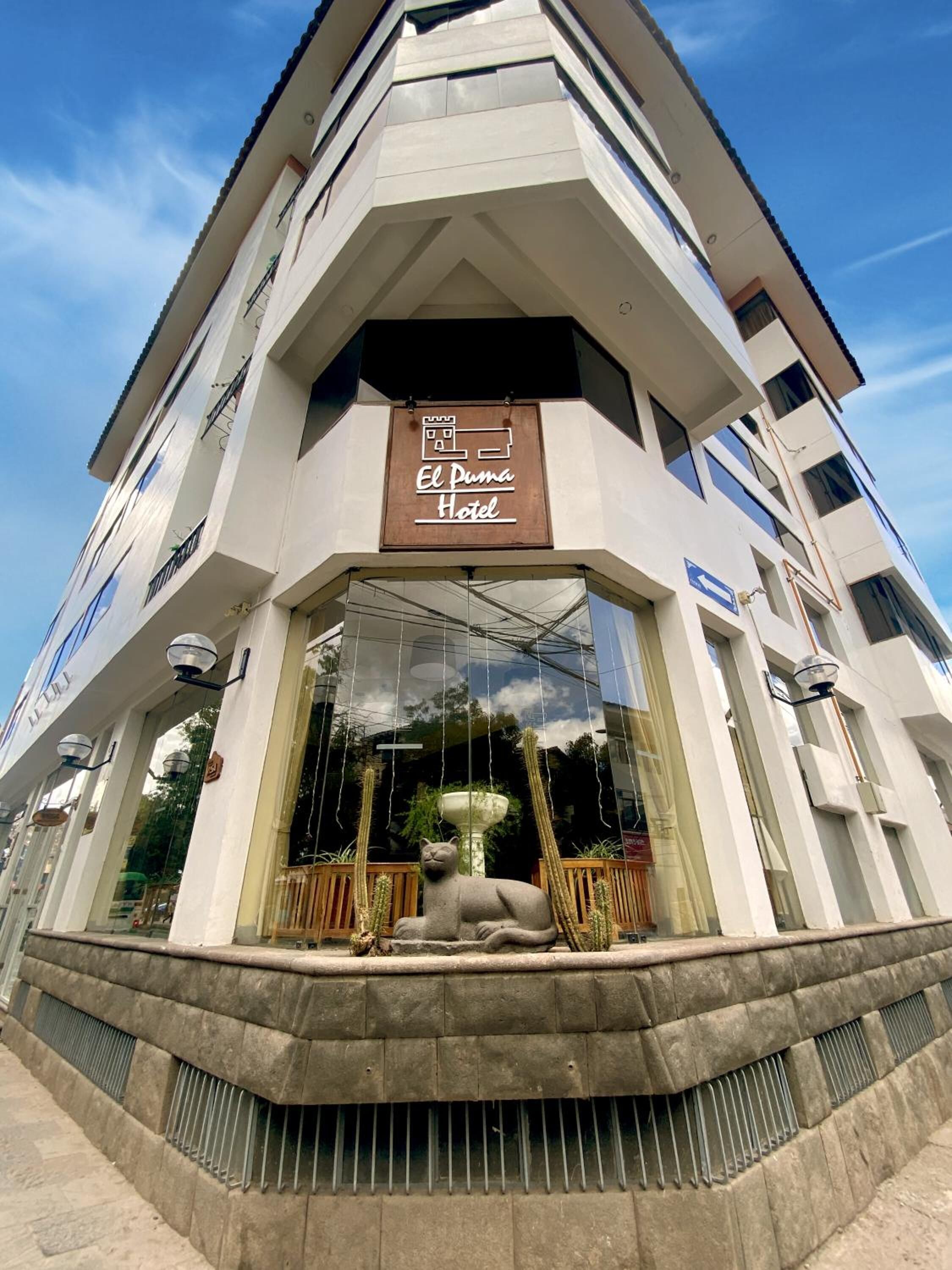 Um hotel chamado "El Puma Hotel", com design moderno, uma grande entrada de vidro e uma escultura de cacto na frente.
